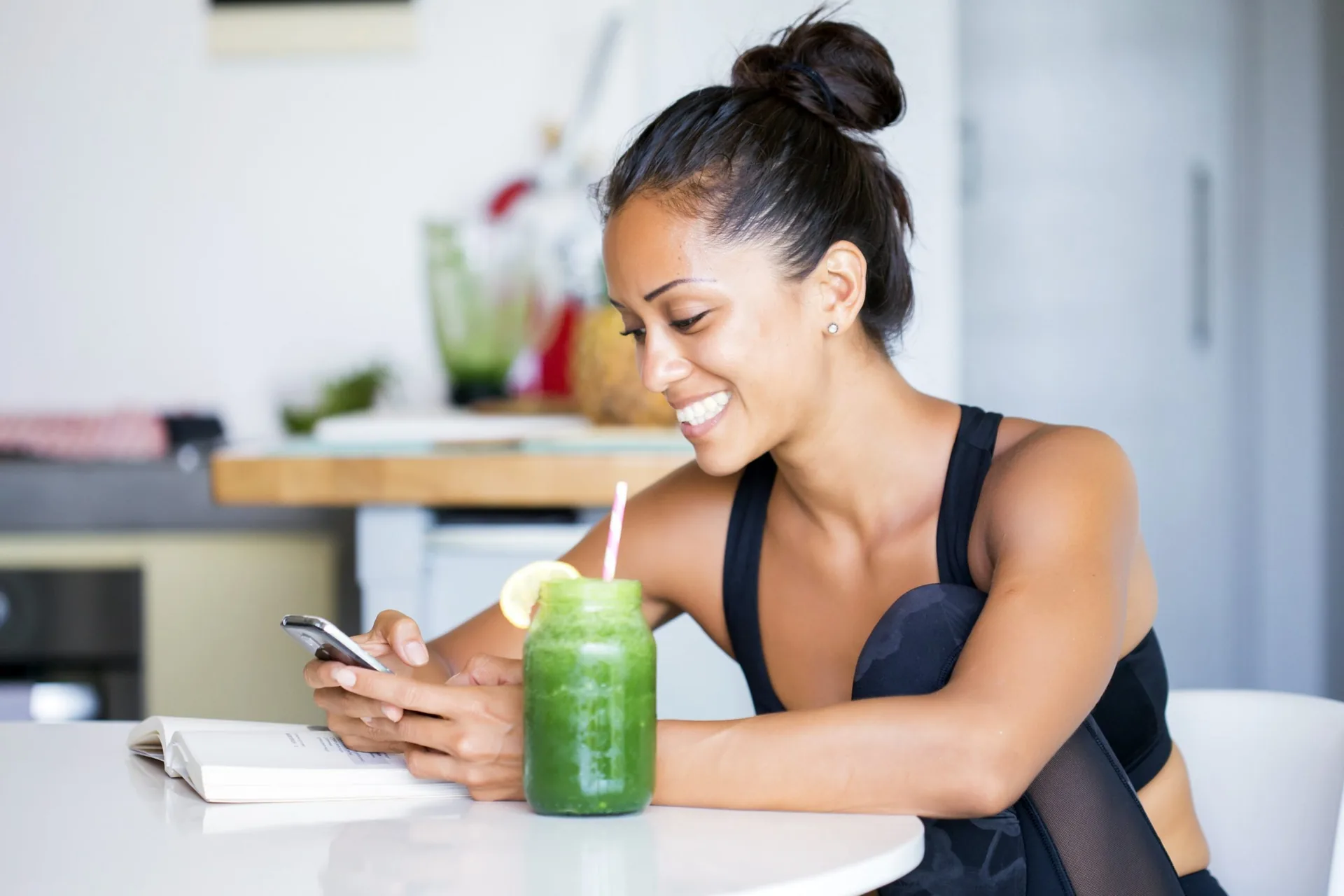 Mulher sorrindo, de roupa esportiva, sentada à mesa da cozinha com um copo de suco verde e o celular na mão